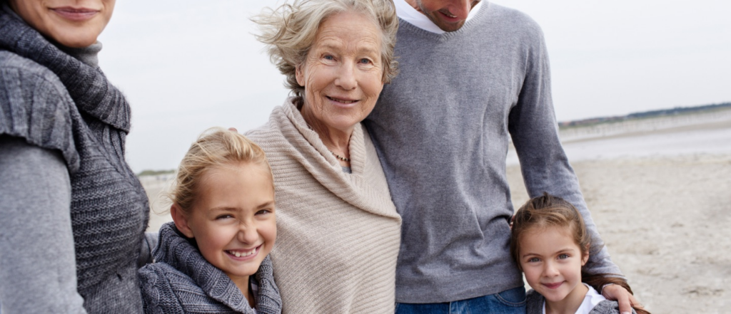 A multigenerational family smiling together on a beach, dressed in cozy sweaters, with a serene shoreline in the background.