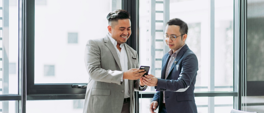 Two men in business attire smiling and discussing something on a smartphone in a bright office space with large windows in the background.