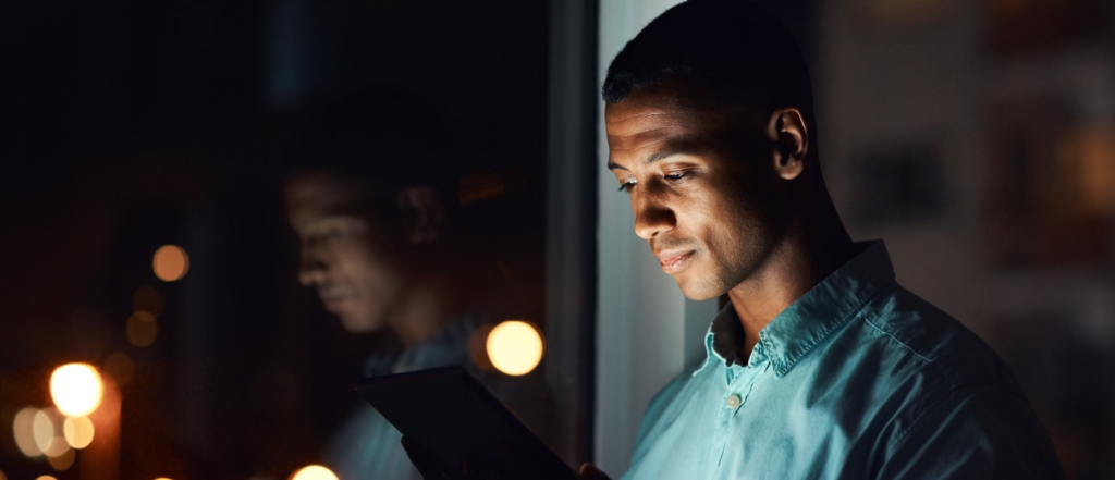 A man in a dimly lit environment reading from a tablet, with his reflection visible in a nearby window and warm lights glowing in the background.