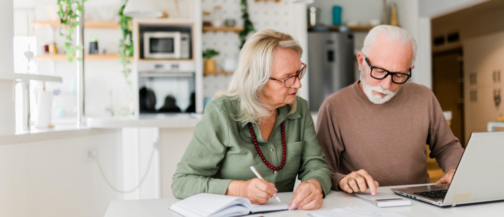 An older couple sitting together at a kitchen table, reviewing documents and working on a laptop, with a modern kitchen in the background.