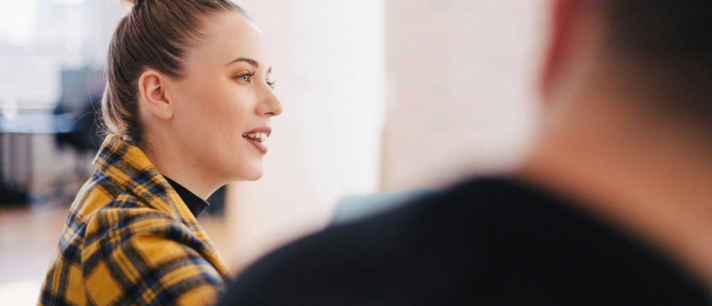 A woman with her hair in a bun wearing a yellow plaid jacket speaking in a bright and modern office environment, with a blurred figure in the foreground.