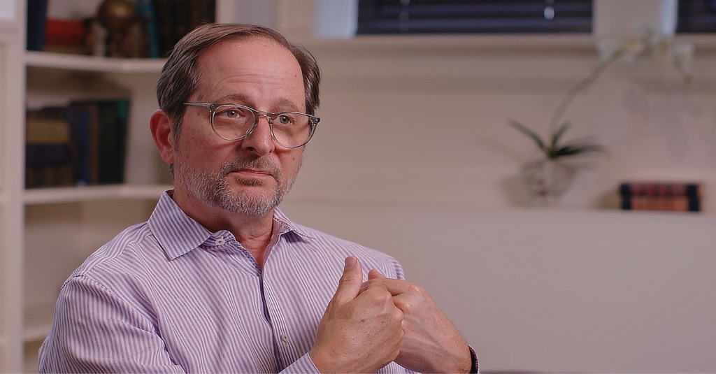 A man wearing glasses and a striped shirt gestures with his hands while speaking, seated in a room with bookshelves and a potted plant in the background.