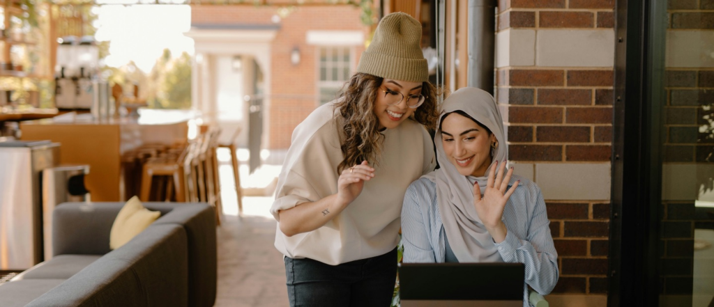 Two women smiling and waving at a laptop screen while sitting in an outdoor café setting, surrounded by warm brick walls and comfortable seating.