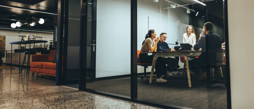 A team of professionals having a lively discussion around a table in a modern glass-walled conference room, with a stylish lounge area visible outside.