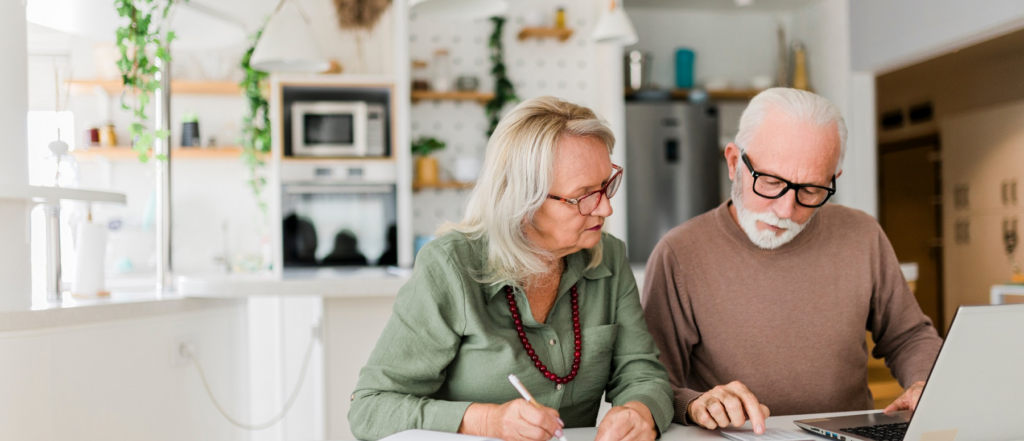 An older couple seated at a kitchen table reviewing documents together, with a laptop open in front of them in a bright, modern kitchen setting.