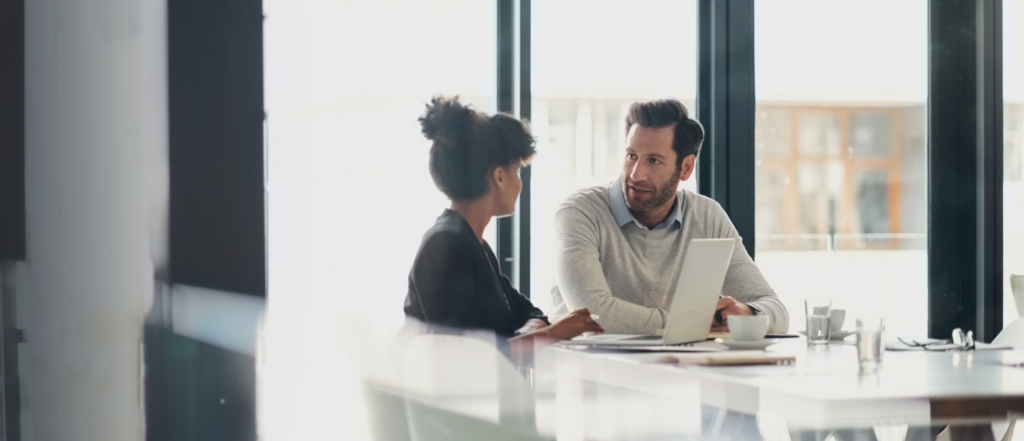 Two professionals seated at a bright, modern table, engaged in a thoughtful discussion with a laptop and coffee cups in front of them.